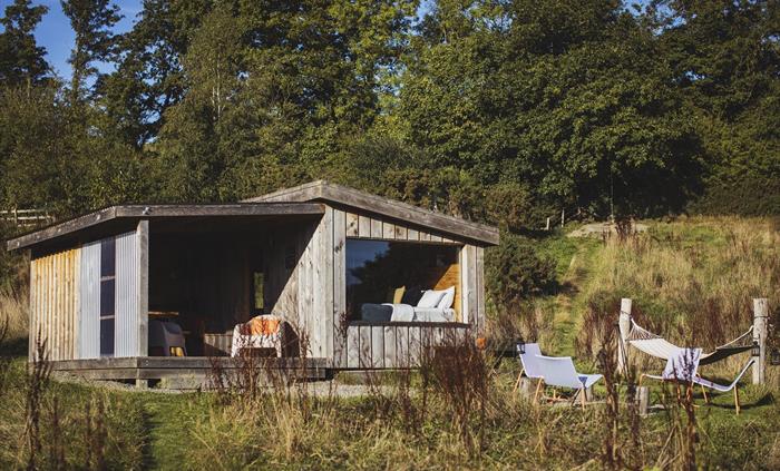 Wood cabin set in a meadow surrounded by trees.  There is large window at the front of the cabin with a bed made up inside. Outdoors is a campfire set